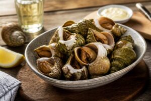 Bulots vivants cuits au court-bouillon servis dans un bol blanc avec une mayonnaise maison, vue de dessus sur fond de bois clair